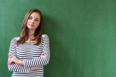 年轻自信的女中学生站在黑板前的教室里, 双臂交叉, 看着镜头。腰上画像