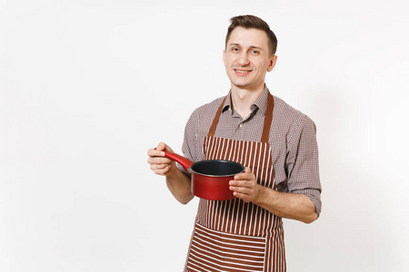 年轻的微笑的人厨师或服务员在条纹棕色围裙, 衬衣拿着红色空的 stewpan, 平底锅或罐在白色背景隔绝。男管家或 housew
