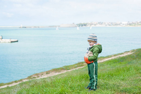 可爱体贴的小男孩穿着围巾夹克和帽子在绿色的草坡反对海上与浮动游艇在春季