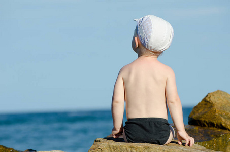 他背对岩石坐在海边游泳裤 蓝蓝的天空 文本空间的小男孩