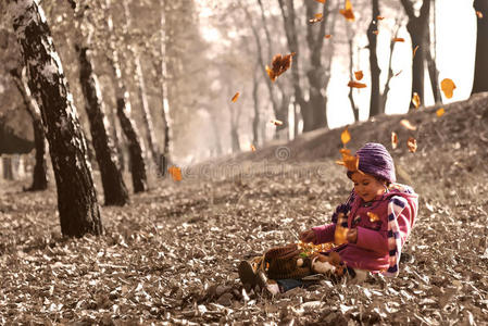 可爱的女孩坐在秋天的落叶上，树叶飘落，玩弄着洋娃娃