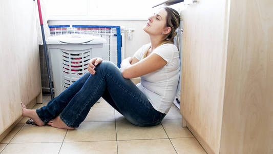 心烦的年轻主妇坐在地板上洗衣和靠在洗衣机