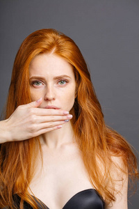 红头发的女人用手捂着嘴巴, 在灰色背景下看着相机