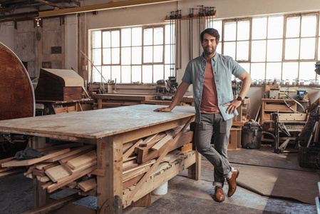 一个自信的年轻木工站在他的木工车间里满是木头的工作台旁边的肖像