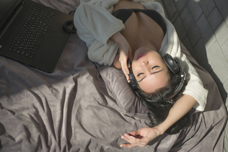 美丽的女人在黑色内衣和白色浴衣听音乐通过耳机, 而躺在床上与笔记本电脑