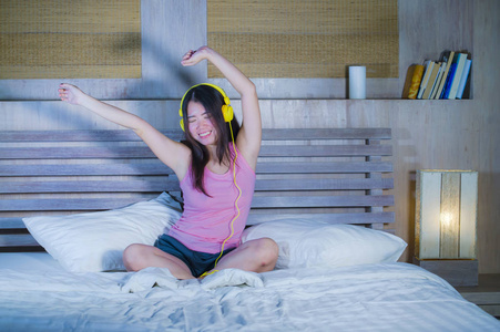 年轻的迷人和快乐的亚洲中国妇女与黄色耳机听音乐在家里的床上的手机上微笑着快乐