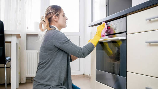 年轻女子用黄色橡胶手套洗烤箱玻璃门