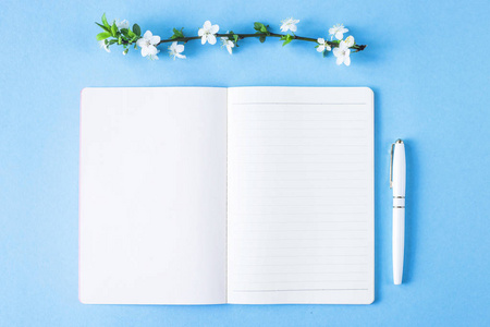 一个打开的空白笔记本, 在蓝色背景上有一支白色钢笔和盛开的树枝。春季工作场所概念