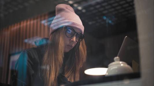 一个时尚的时髦女人在眼镜和帽子坐在咖啡馆和打字与笔记本电脑。通过窗口查看
