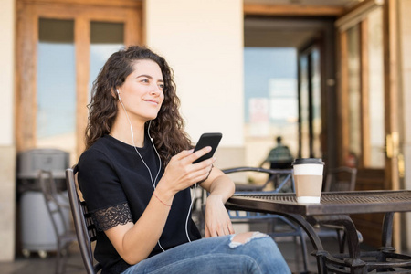美丽的年轻女子在户外咖啡馆放松, 从她的手机听音乐