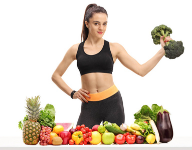 健身妇女与西兰花哑铃在桌子后面的水果和蔬菜隔离在白色背景