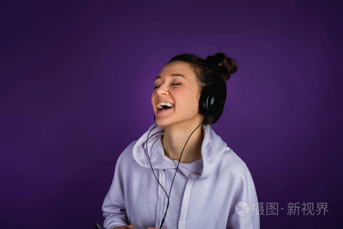 的年轻女孩与白色的牙齿听音乐和在一个紫色的背景运动衫的耳机唱歌