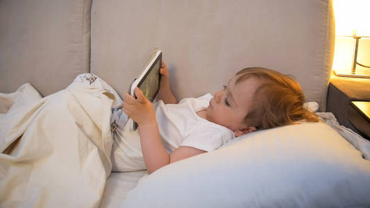 可爱的蹒跚学步的男孩在床上看卡通片的肖像