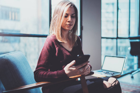 手持现代手机的金发青年女人。女孩指着手指在阳光明媚的 coworking 地方的空触摸移动屏幕。横向模糊背景