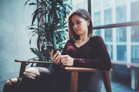 手持现代手机的金发青年女人。女孩指着手指在阳光明媚的 coworking 地方的空触摸移动屏幕。横向模糊背景