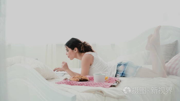 年轻美丽的女人躺在床上和使用笔记本电脑。女孩上网冲浪的在早晨吃早饭