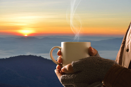 手在手套拿着咖啡杯子山和太阳的背景