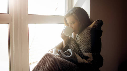 美丽的年轻女子披着格子在橱窗里喝茶