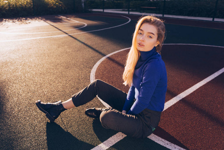 时尚的年轻模特金发女郎在一个蓝色的毛衣摆在一个体育领域在阳光下