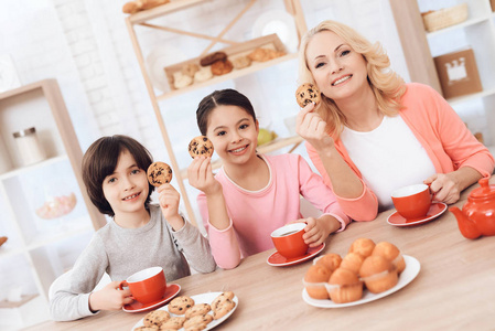 年轻的祖母与孙子和孙女吃曲奇饼和喝茶在厨房的红色杯子
