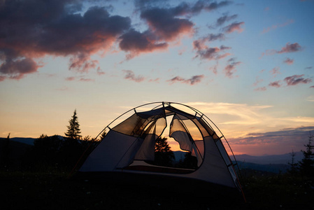 帐篷孤零零地矗立在草地上, 上面是一座小山上的野花, 有云的美丽夜空。杉木和大山背景下日落露营的魔幻景观