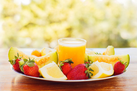 一杯新鲜的橙汁在金属盘子上, 上面有一束鲜艳的水果。草莓, 瓜, 柚子在花园的背景上的老木桌上的街道上