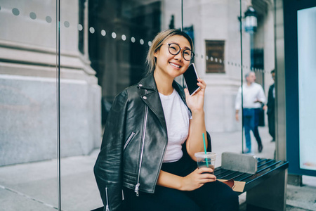微笑的亚洲女孩在眼镜和凉快的穿戴讲电话坐在公共汽车站长凳户外在市中心, 开朗的中国年轻妇女打电话给朋友使用漫游国际连接