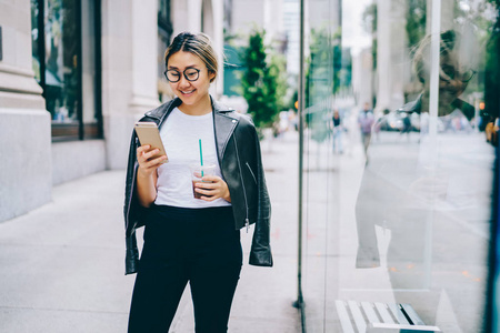 微笑的亚洲妇女在时髦穿戴步行在城市街道与朋友在网络上聊天通过移动, 正面女性中国游客浏览信息关于市中心咖啡馆的电话