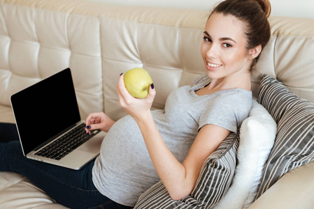快乐怀孕的年轻女子在沙发上使用空白屏幕笔记本电脑