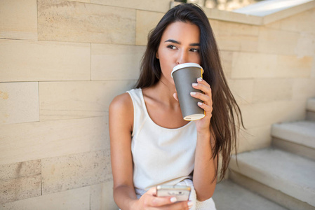 美丽的肖像年轻迷人的黑发妇女喝咖啡或茶, 坐在公园的台阶上, 手机短信。看着一边思考着