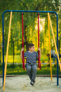 一个穿着条纹 t 恤的小男孩正在操场上玩，在秋千上摇摆。春天，阳光明媚的天气
