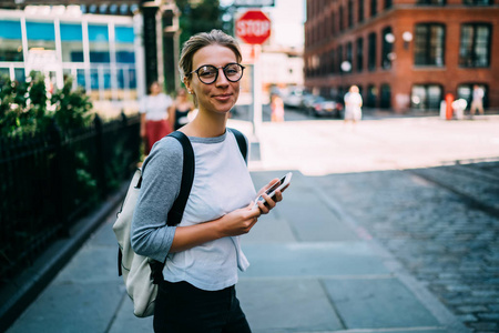 愉快的女性的肖像在城市街道上漫步手持智能手机发送邮件给朋友, 微笑的时髦女孩看着相机花周末旅行检查位置