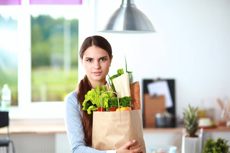 年轻女子捧着蔬菜杂货店购物袋。站在厨房