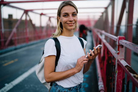 半长的微笑的时髦女孩的肖像站在城市背景与智能手机, 积极的年轻女子在衬衫与复制空间的标签发送消息在城市的街头散步