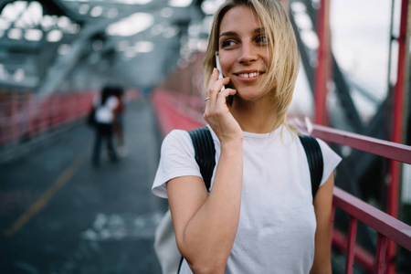 微笑的年轻妇女在 t恤与拷贝空间为标签或牌子名字谈论在移动电话步行, 时髦的女孩满意为拨打电话在漫游有电话谈话的关税