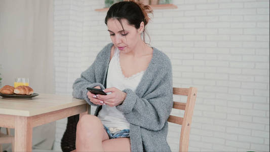 美丽的女人坐在一个舒适的公寓和上网冲浪。黑发的女孩使用智能手机和喝咖啡