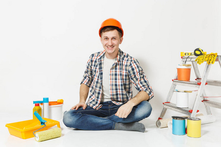 微笑的人在橙色防护头盔坐在地板上的翻新公寓室的设备, 在白色背景隔离。墙纸胶水配件, 喷漆工具。修复主页概念