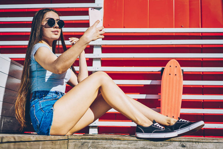 有吸引力的休闲打扮时髦的女孩在时尚的太阳镜制作自拍照片的前相机的现代智能手机设备休息与滑板户外。在蜂窝上拍照的年轻女子