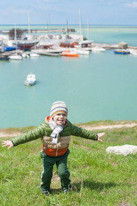 愉快的小男孩穿着温暖的夹克围巾和帽子在绿色斜坡反对海港的背景与小船在冷的春天