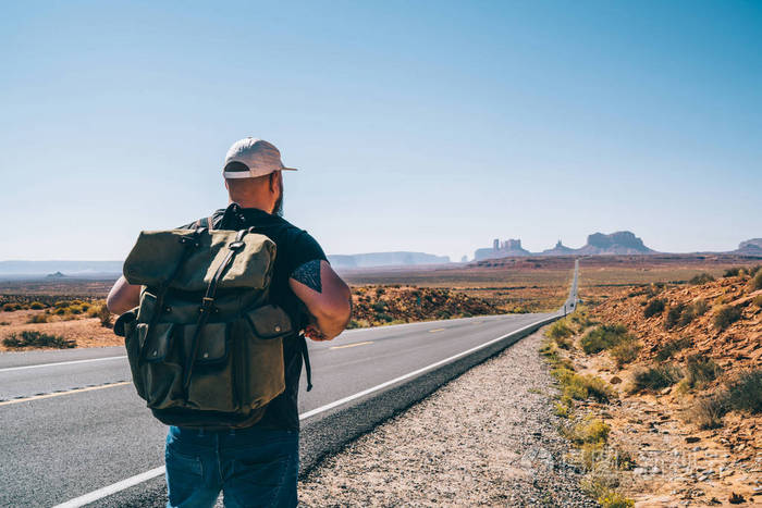 男性游客流浪者的后面看着背包去纪念碑谷在沥青路在狂放的环境时髦的