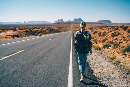 时尚女孩流浪走在西南野生土地上的沥青公路上步行到国家公园步行, 女游客与背包有旅程在亚利桑那州搭便车途中