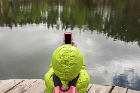 女性旅行者坐在附近一个美丽的湖，在一片松树林和拍照手机上的木码头上