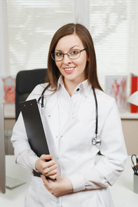 年轻微笑的女医生站在办公桌前与剪贴板和医疗文件在光办公室在医院。妇女在医疗礼服, 听诊器在咨询室。医疗保健, 医学理念