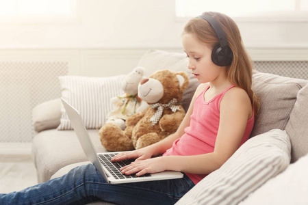 可爱的小女孩在笔记本电脑上做作业, 坐在沙发上戴着耳机