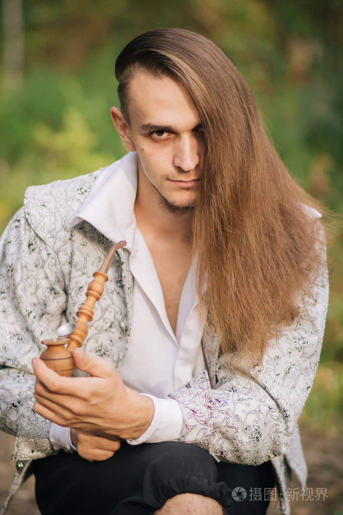 性感长发男子与烟斗在他的手在树林里穿着一件漂亮的古老的夹克上面有