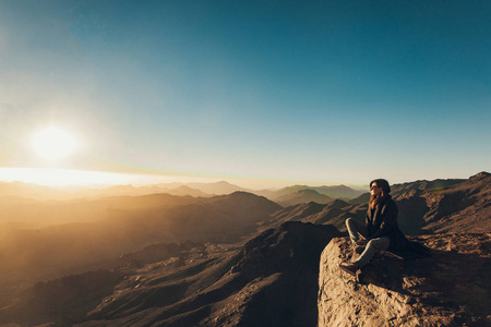 女人坐在西奈悬崖边上, 反对日出的背景。
