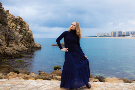 一个美丽的金发女孩在海边是悲哀的多云阴雨天气, 穿着一条长蓝裙, 西班牙海岸