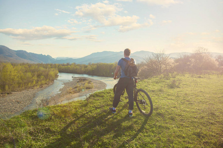 男孩骑自行车站在山上, 看着河在日落的概念为旅客。风景从旅行者的后面在背景山