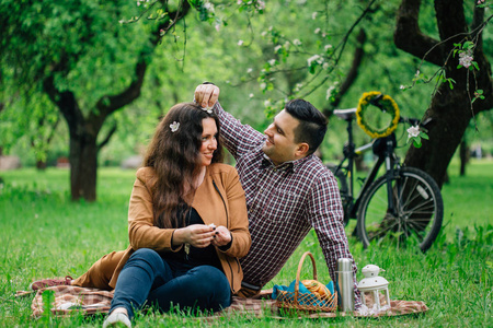 年轻的时尚幸福情侣在盛开的花园和自行车后面野餐。男人把花放在女朋友的头发上