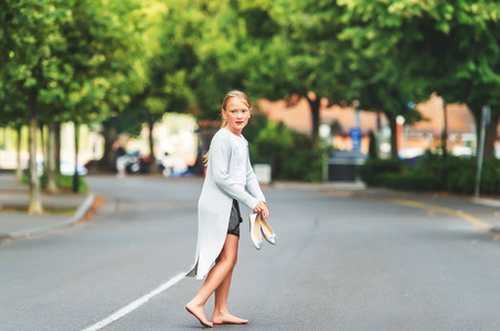 赤脚小女孩走在街上，戴着长灰色羊毛衫，持有银弓芭蕾舞鞋